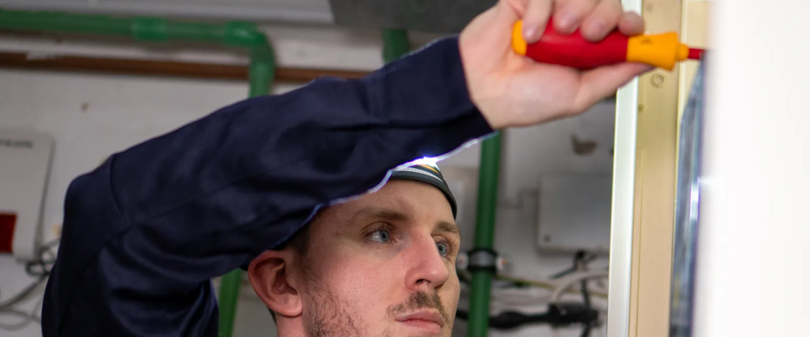 Techniker in dunkelblauer Arbeitskleidung mit Stirnlampe schraubt mit einem isolierten Schraubendreher an einem Elektroverteiler; im Hintergrund grüne Rohrleitungen und Kabel eines Technikraums.